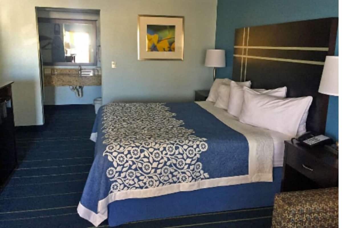 A hotel room with a bed, a desk, a chair, a lamp, a painting on the wall, and a bathroom visible through an open door.
