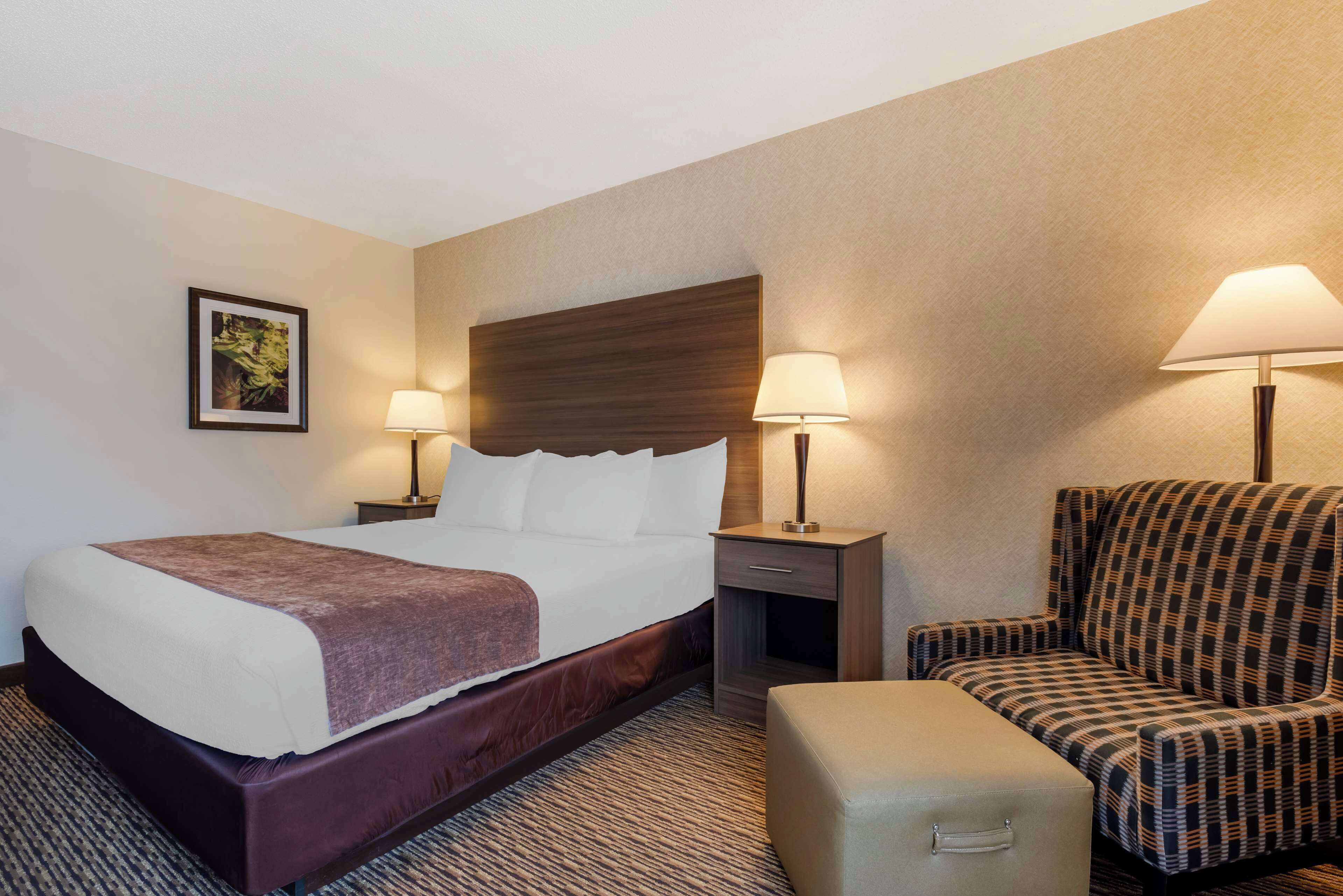 Habitación de hotel con una cama grande, dos mesitas de noche con lámparas, una silla y un pequeño reposapiés.