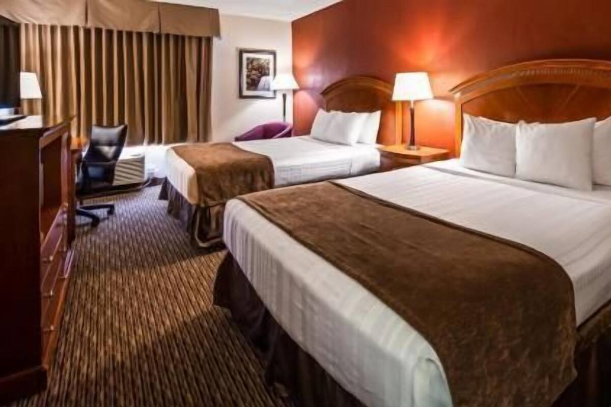 A hotel room with two beds, a desk, a chair, and a lamp.