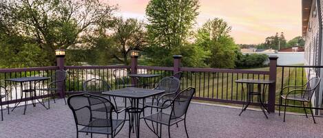 An outdoor patio with tables and chairs, a railing, and a view of trees and a building in the background.