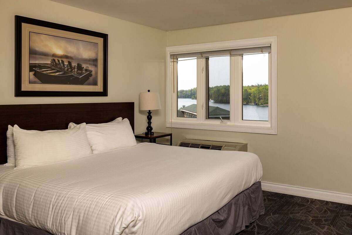 A hotel room with a large bed, a framed picture on the wall, a window with a view of a lake, and a nightstand with a lamp.