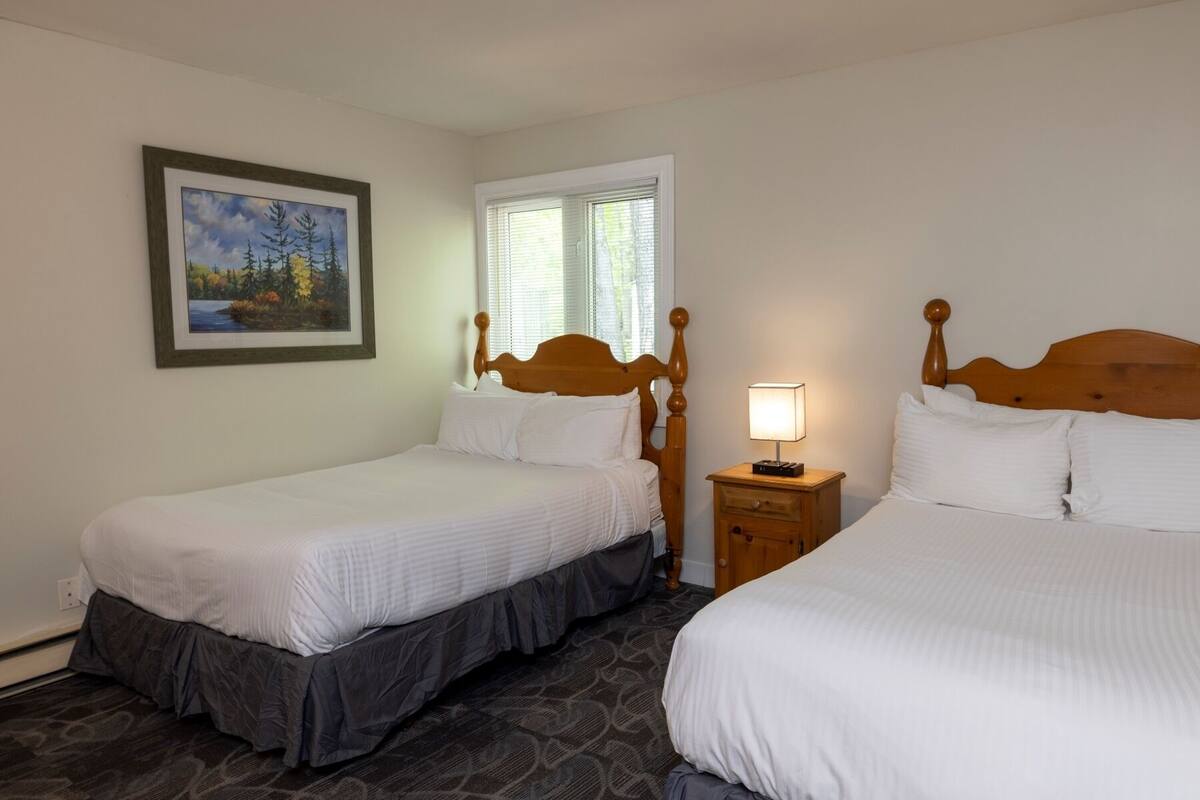 A hotel room with two single beds, a nightstand with a lamp, a painting on the wall, and a window with blinds.