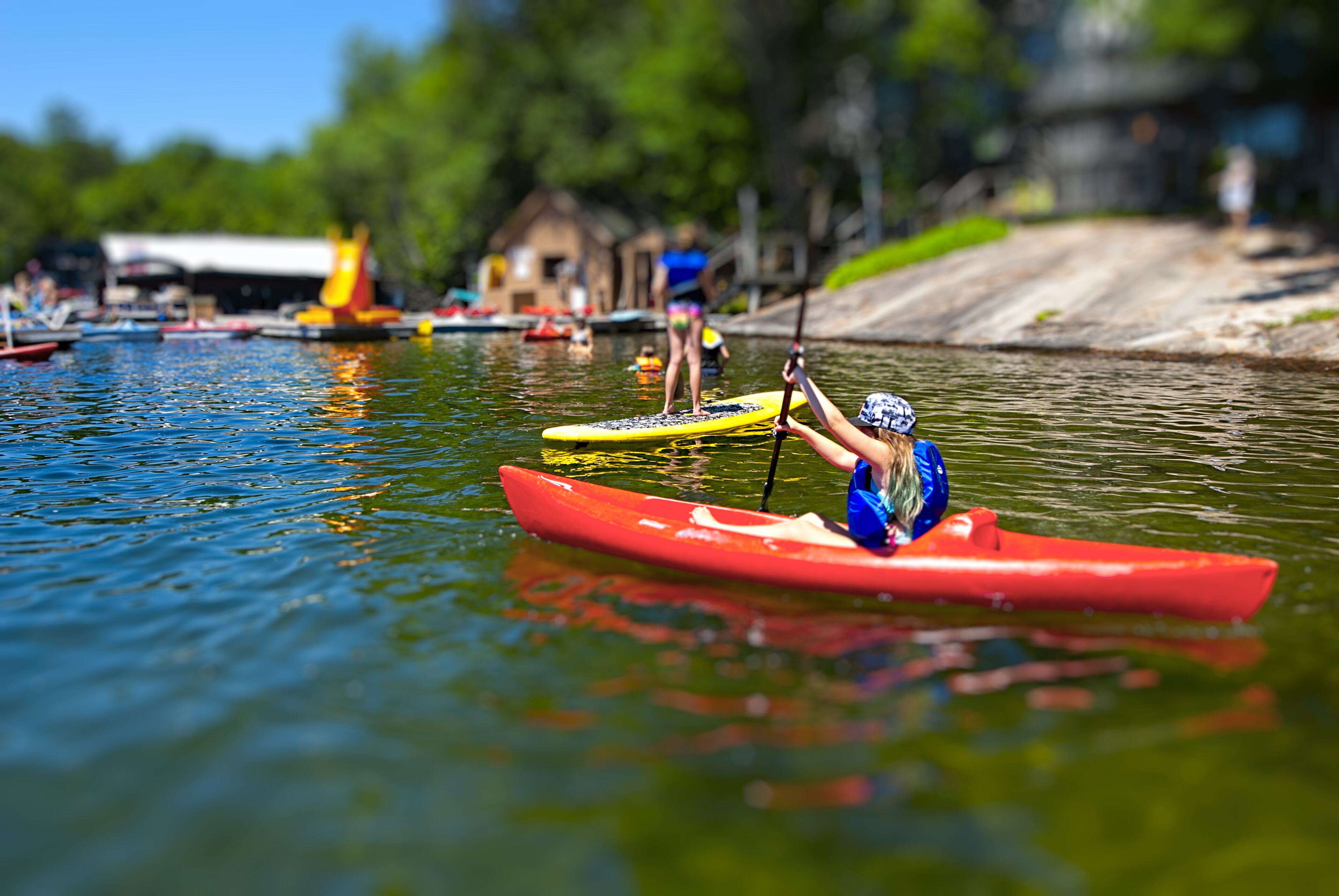 A person in a red kayak on a calm body of water, with other kayaks and people in the background.