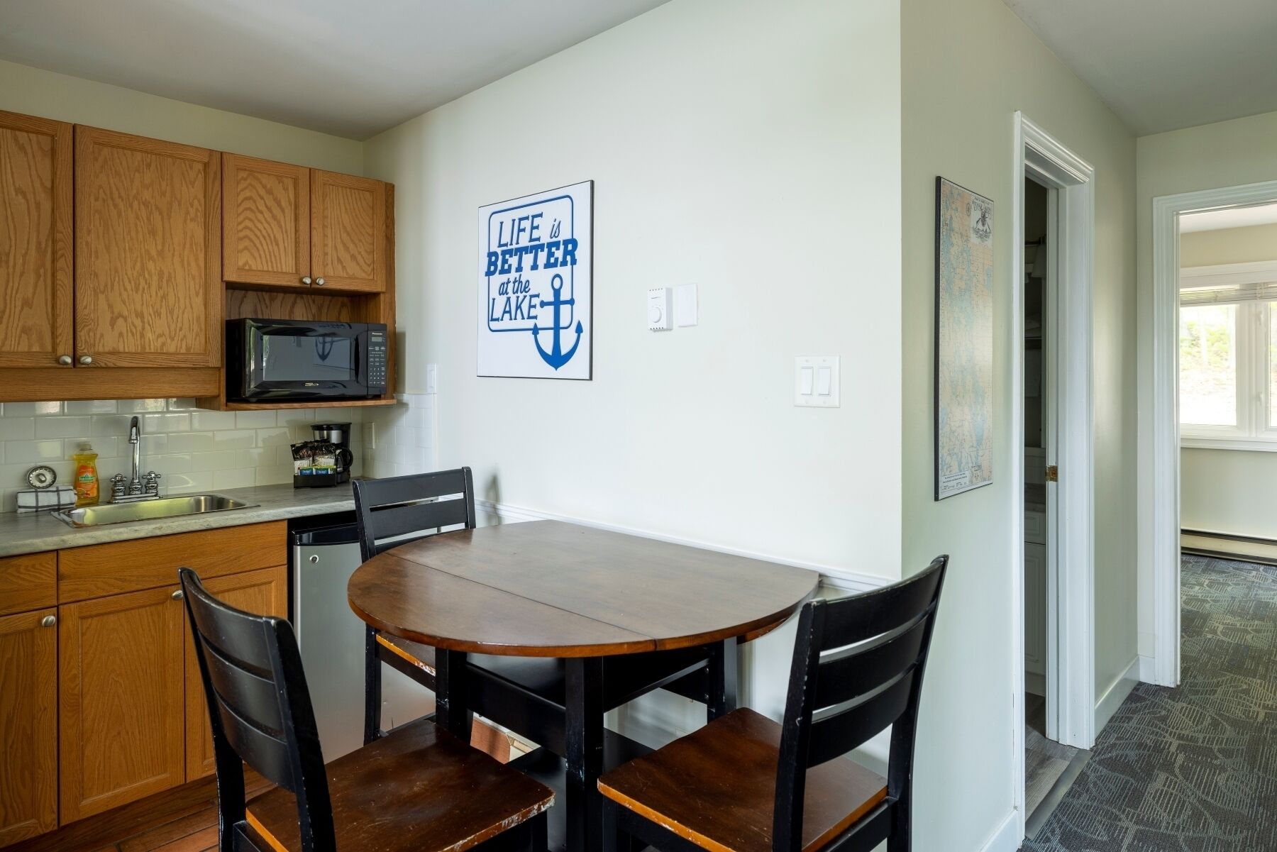Una cocina compacta con armarios de madera, un microondas, un lavabo y una mesa de comedor con sillas. Una frase motivacional está expuesta en la pared.