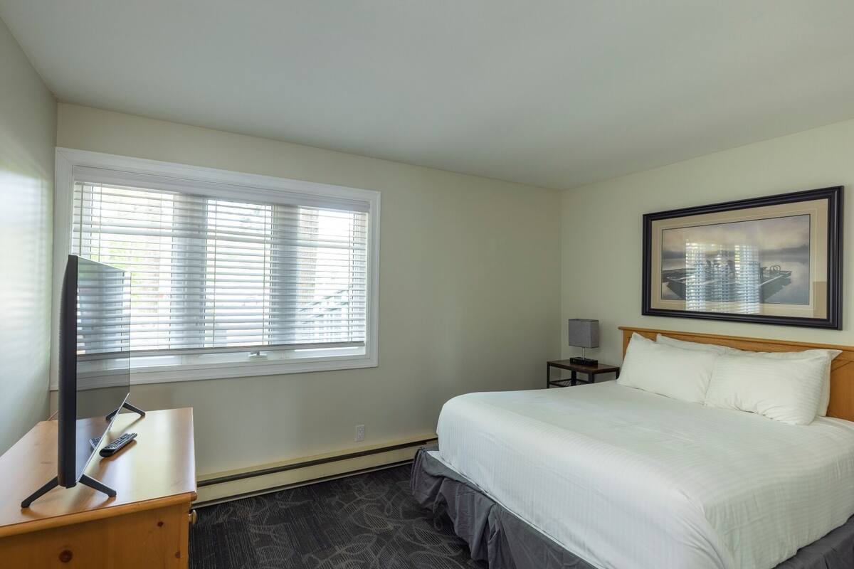 A hotel room with a bed, a desk with a computer, a window with blinds, a nightstand with a lamp, and a framed picture on the wall.