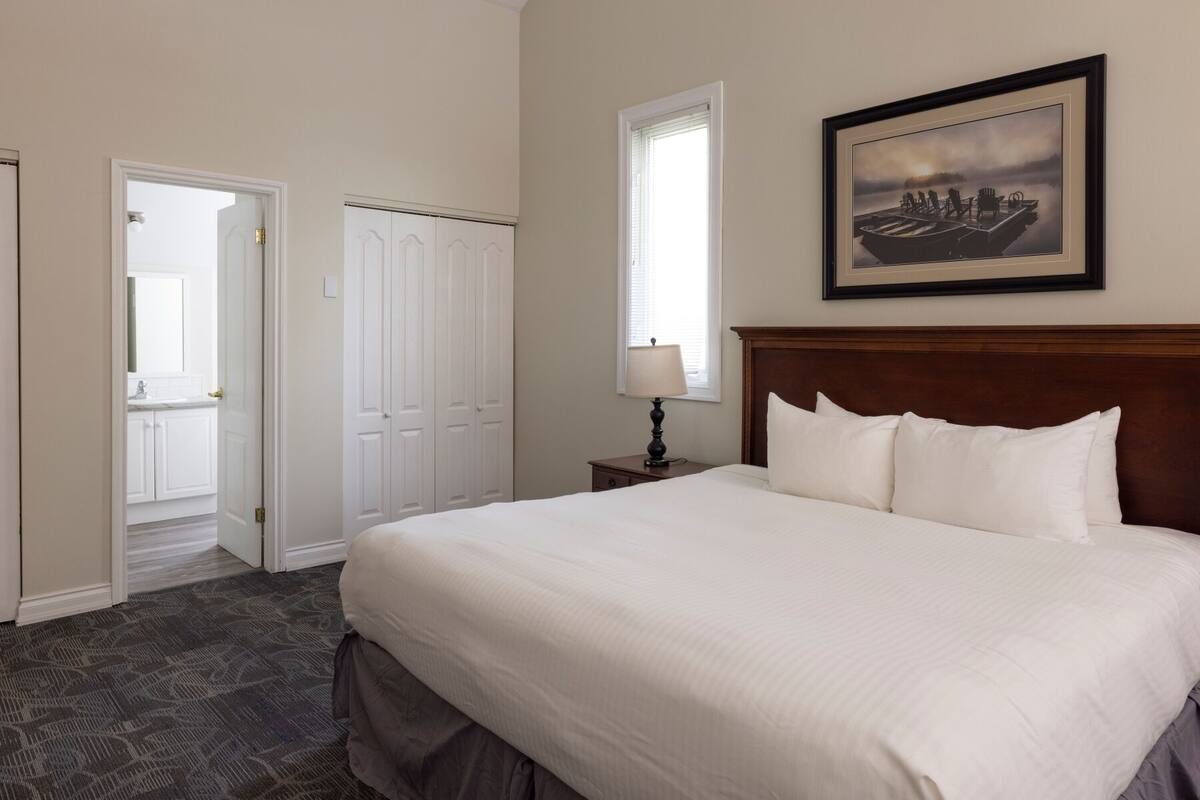 A hotel room with a large bed, a nightstand with a lamp, a framed picture on the wall, and a view of the outside through a window.