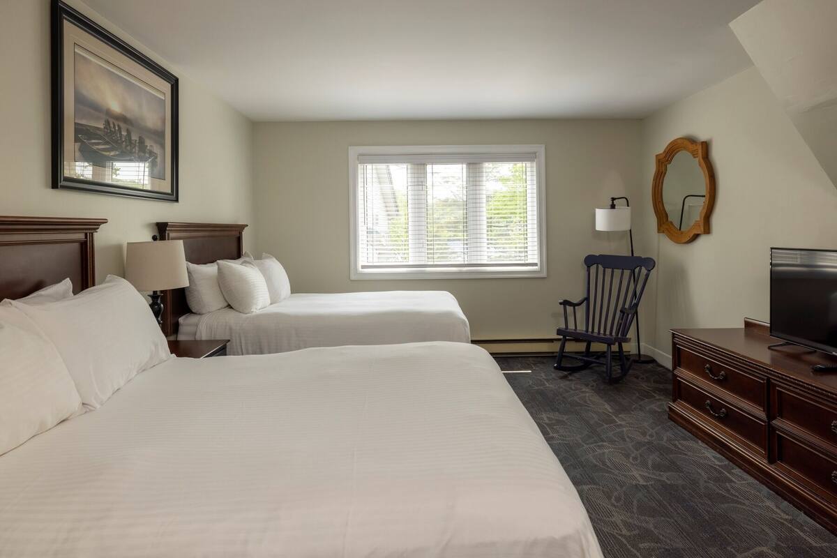 A hotel room with two beds, a desk, a chair, a TV, and a window with blinds.