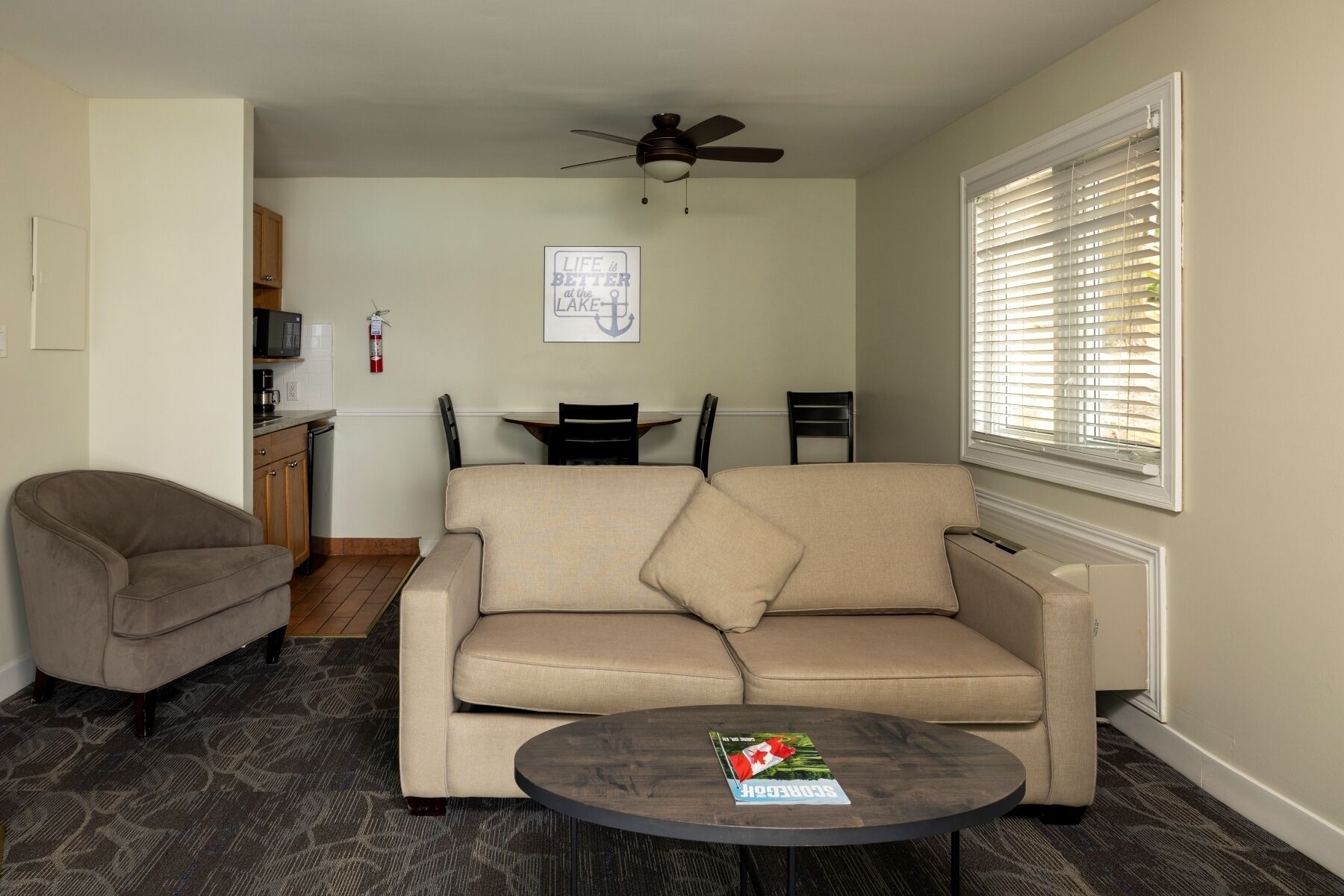 A living room with a beige sofa, a round coffee table, and a ceiling fan. There is a dining area with a table and chairs, and a kitchen area with cabinets and a microwave.