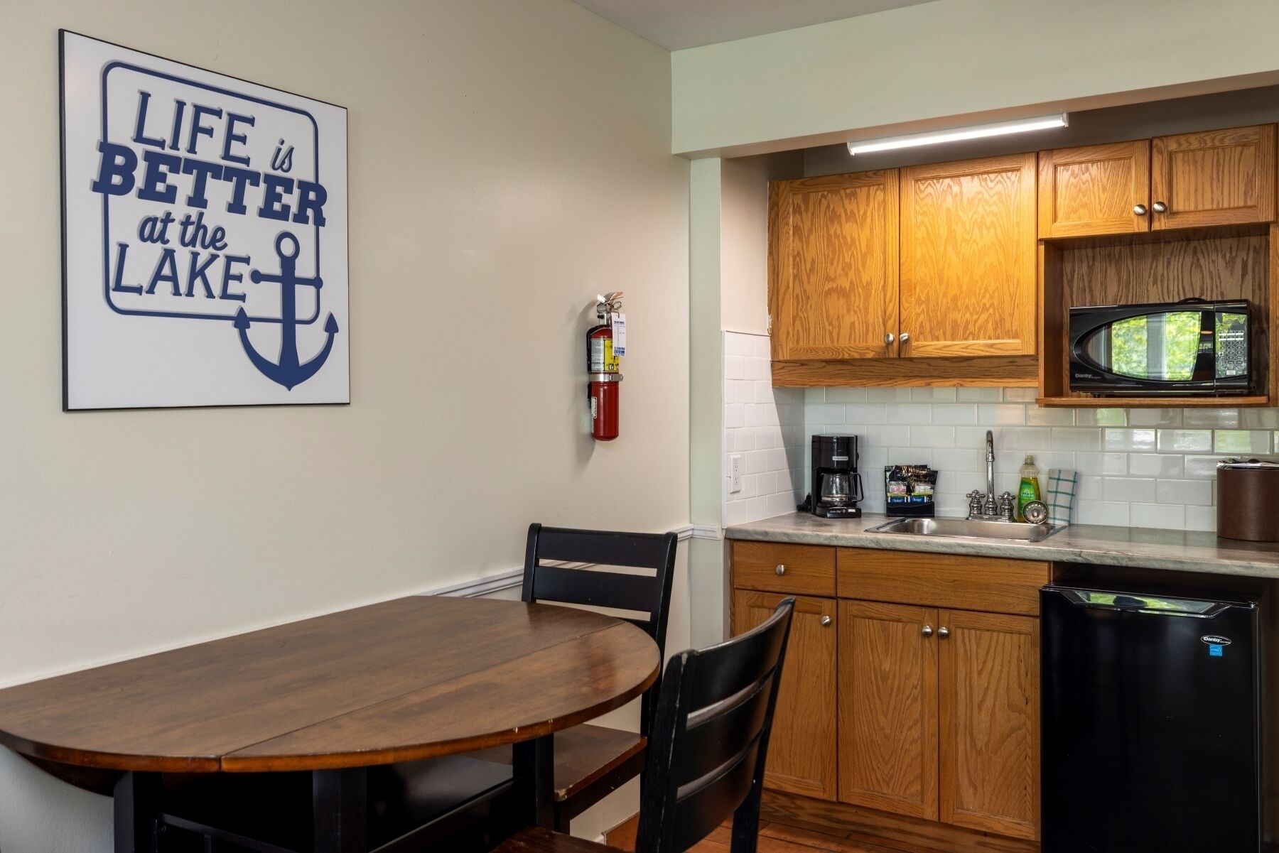 A kitchen with wooden cabinets, a microwave, a coffee maker, and a fire extinguisher. There is a dining table with chairs and a sign on the wall.