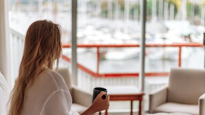 Eine Person sitzt auf einem Bett in einem Hotelzimmer mit einem großen Fenster mit Blick auf einen Yachthafen.
