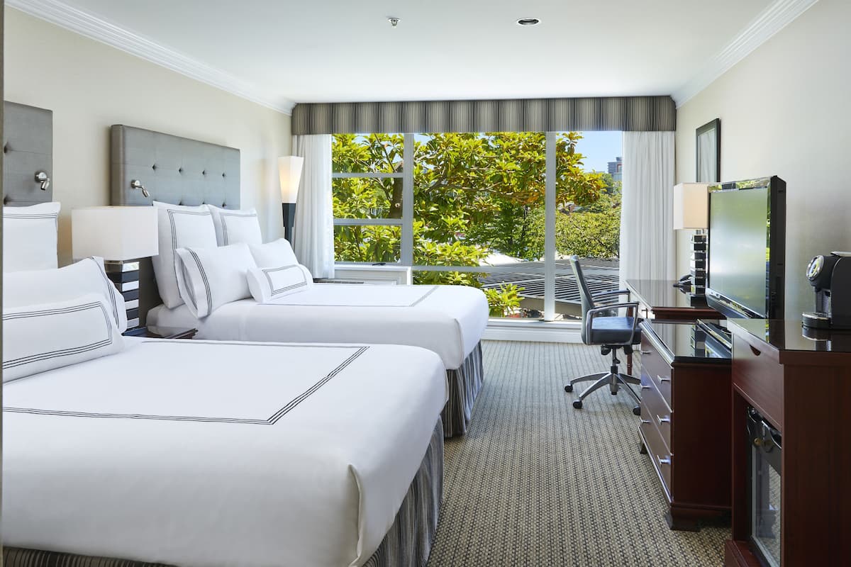 A hotel room with two beds, a desk, a TV, and a large window with a view of trees.