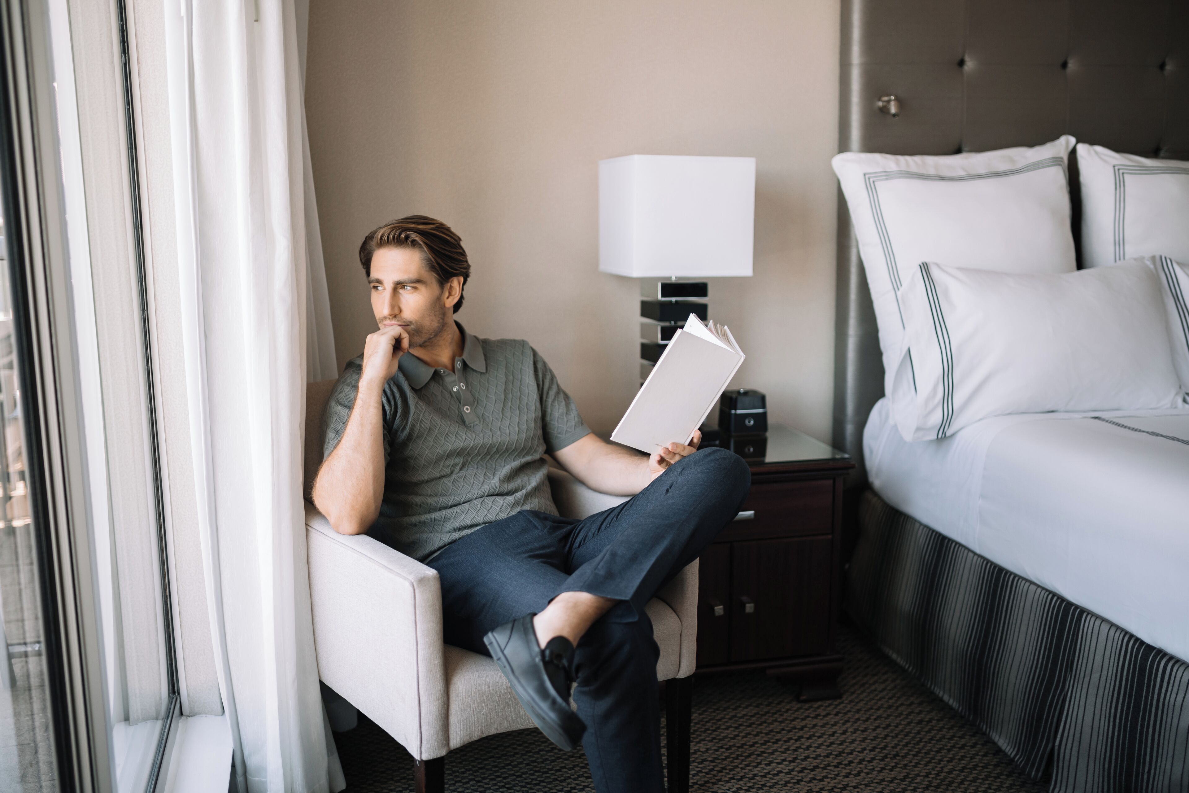 A man sitting in a hotel room, holding a book. The room has a bed with white bedding, a nightstand with a lamp, and a window with curtains.