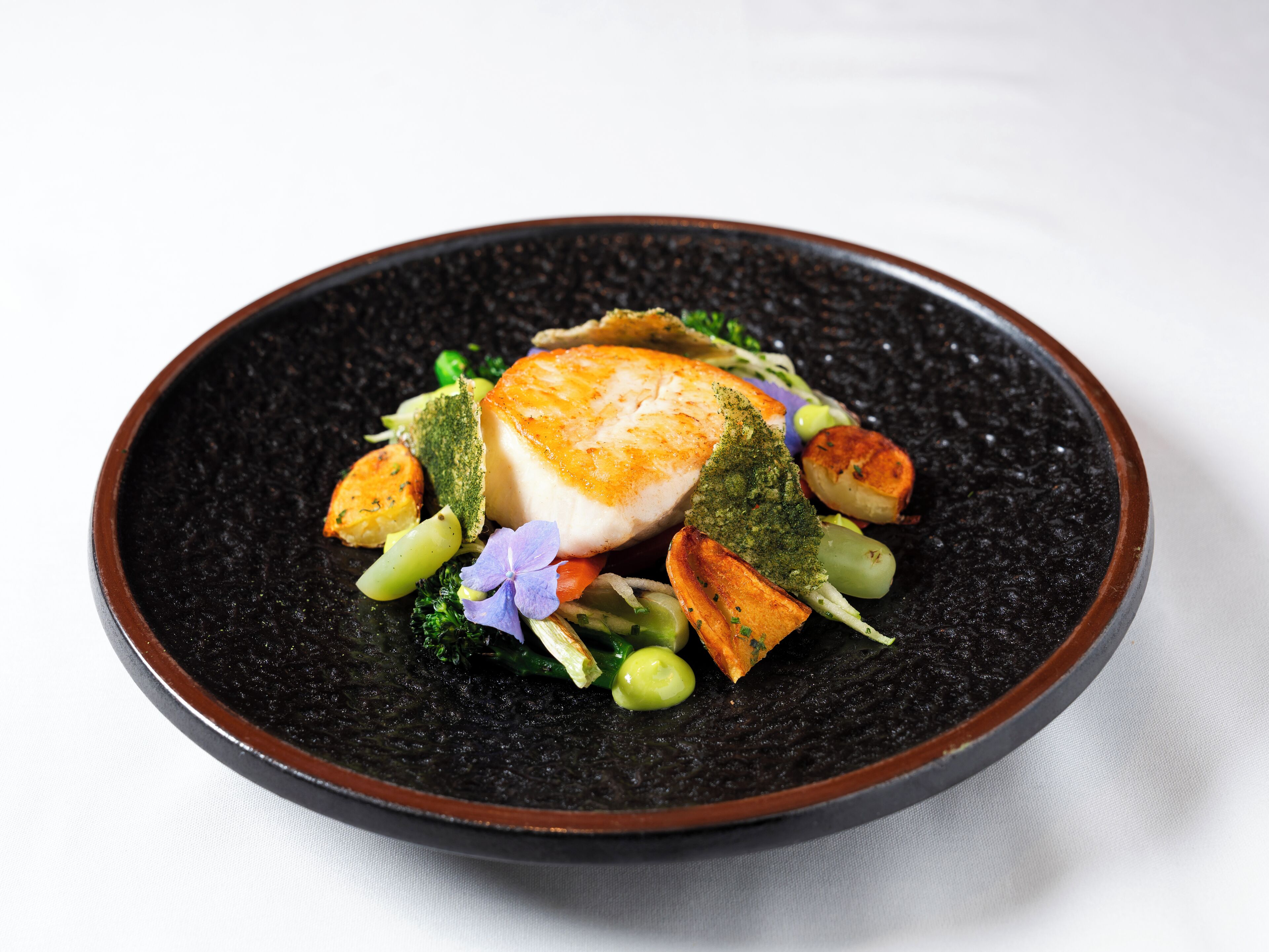 A dish with a piece of fish, vegetables, and a flower on a dark plate.