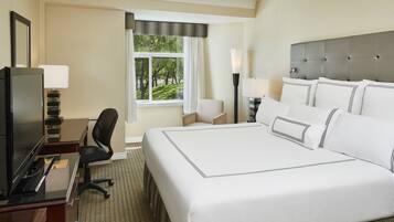 A hotel room with a large bed, a desk with a computer, a chair, a TV, and a window with a view of trees.