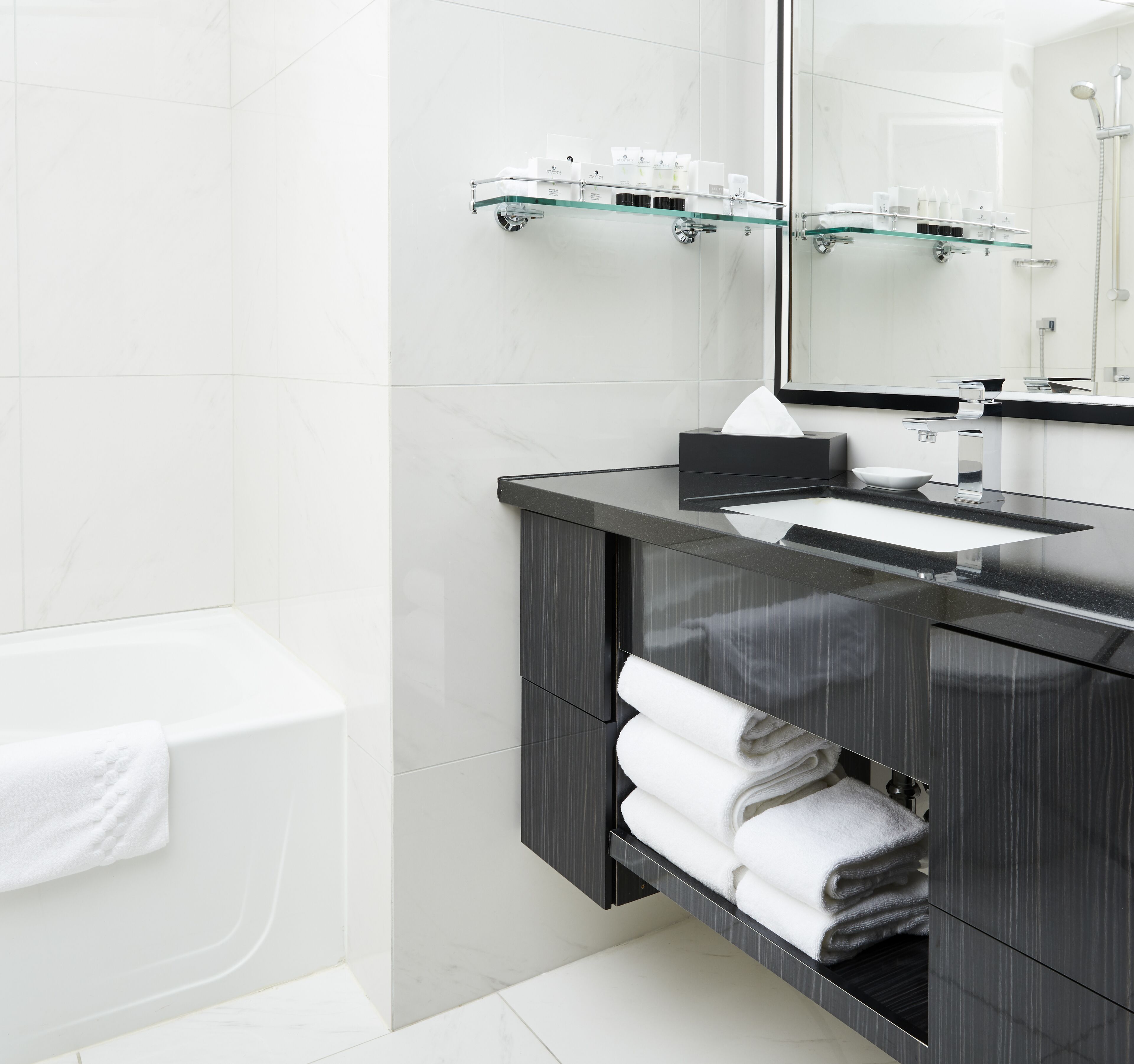 A modern bathroom with a glass shelf holding toiletries, a black vanity with a sink, and a towel rack with folded towels.