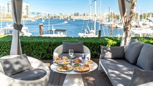 Uma área de refeições ao ar livre com móveis de vime e uma mesa posta, com vista para uma marina com barcos e prédios da cidade ao fundo.