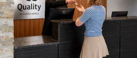 Un mostrador de recepción de hotel con un letrero que dice 'Quality by Choice Hotels', una recepcionista y un huésped interactuando.