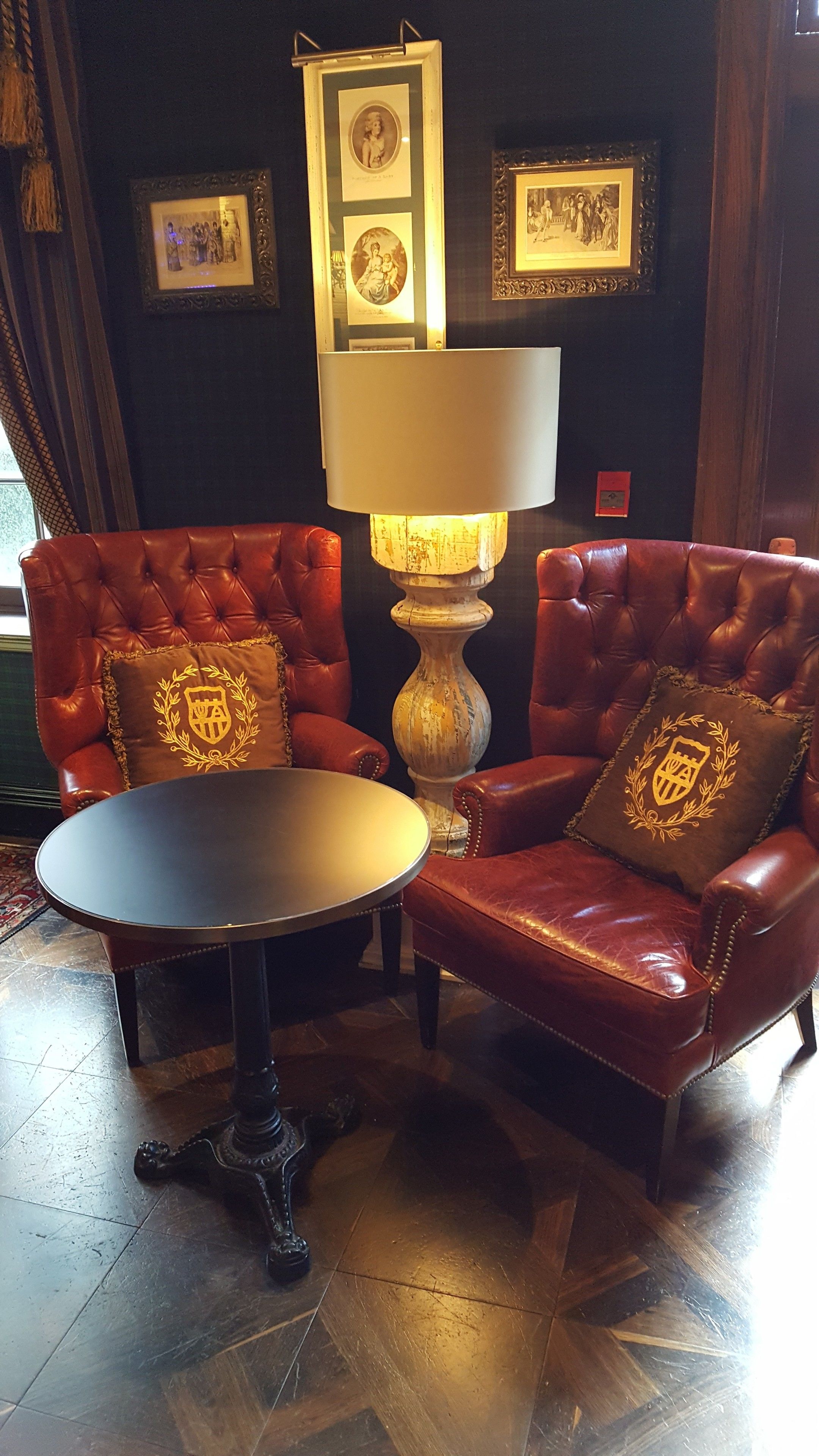A room with a tufted leather armchair, a matching ottoman, and a round table. There is a wall-mounted lamp and framed pictures above the armchair.