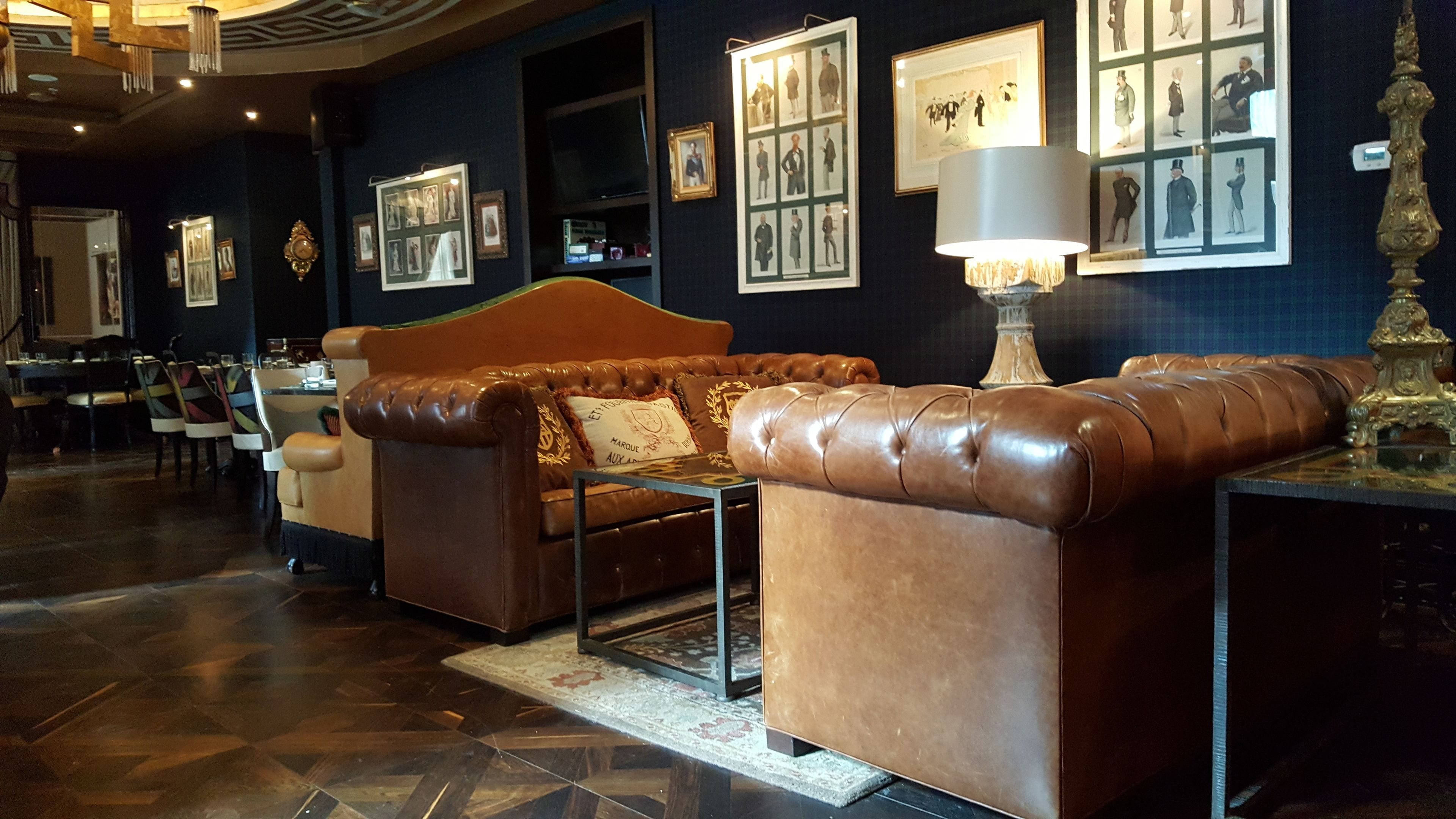 Um lobby com um sofá de couro marrom, uma luminária de mesa, quadros nas paredes e um tapete estampado.