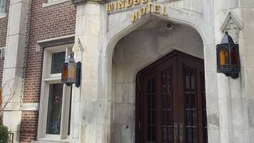 L’entrée de l’hôtel Windsor Arms, avec sa façade en briques, une grande porte en arc de cercle et un mât sur lequel flotte le drapeau américain.
