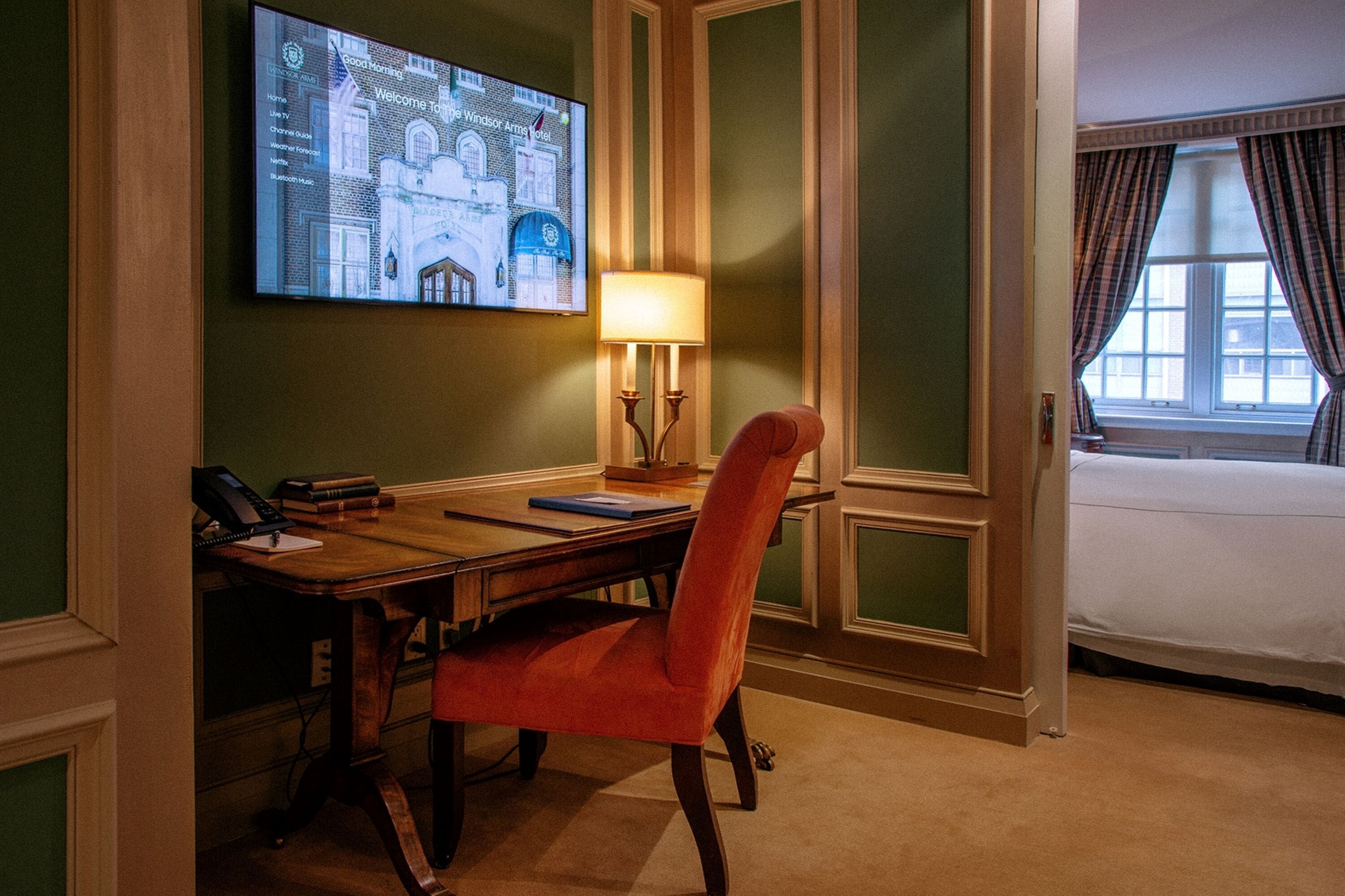 Une chambre d’hôtel avec un bureau en bois, une chaise rouge, une lampe, un lit et un téléviseur affichant un message de bienvenue.