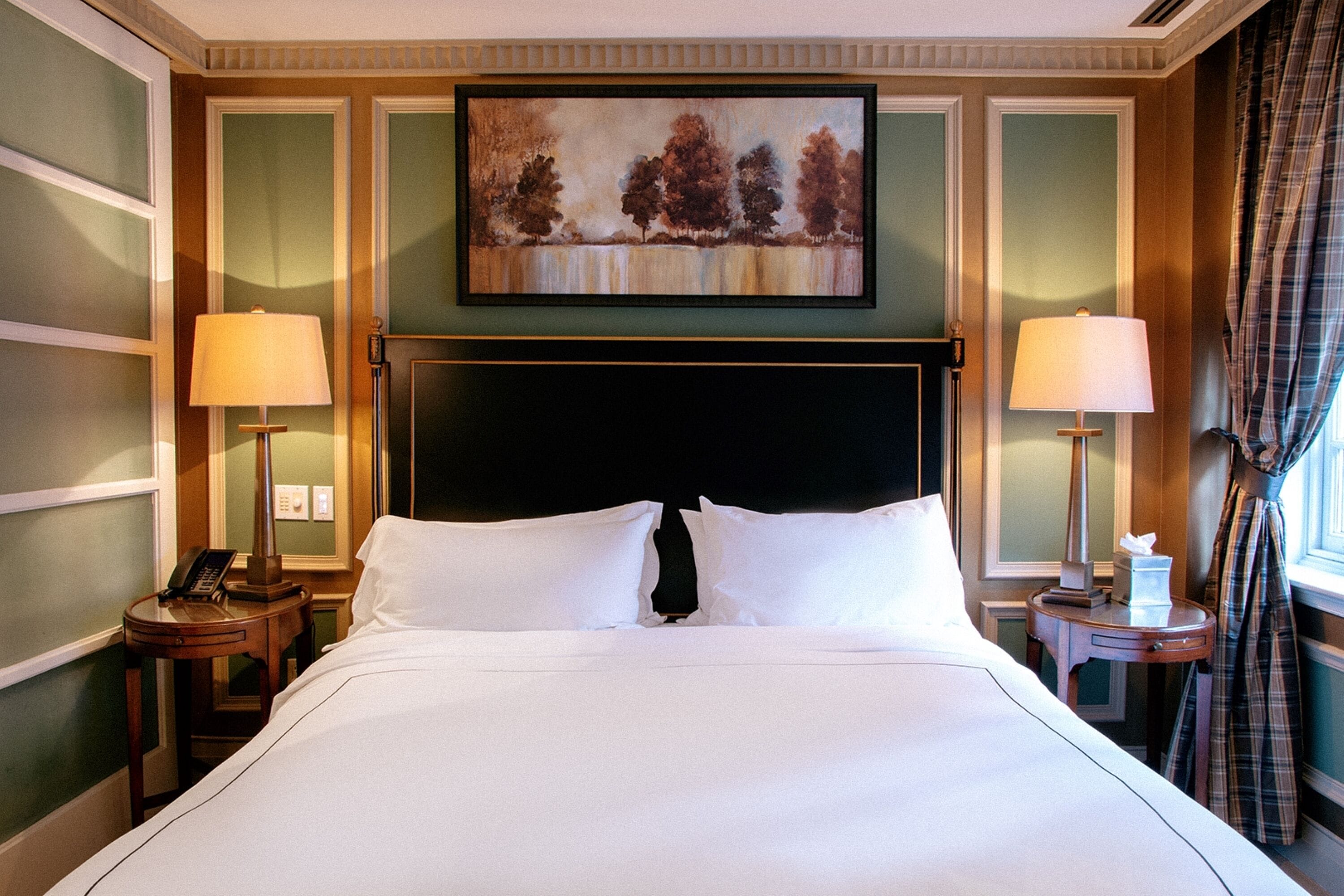 Quarto de hotel com uma cama grande, duas mesas de cabeceira com luminárias, um quadro na parede e uma janela com cortinas.