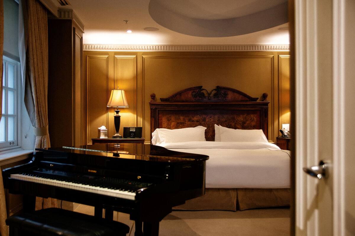 Une chambre d’hôtel avec un grand piano, un lit soigneusement fait et une table de chevet avec une lampe.