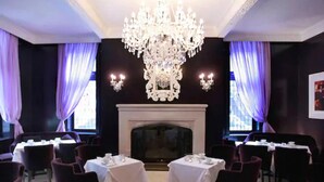 Une salle à manger élégante, avec des tables dressées pour le service, un grand lustre et une cheminée.