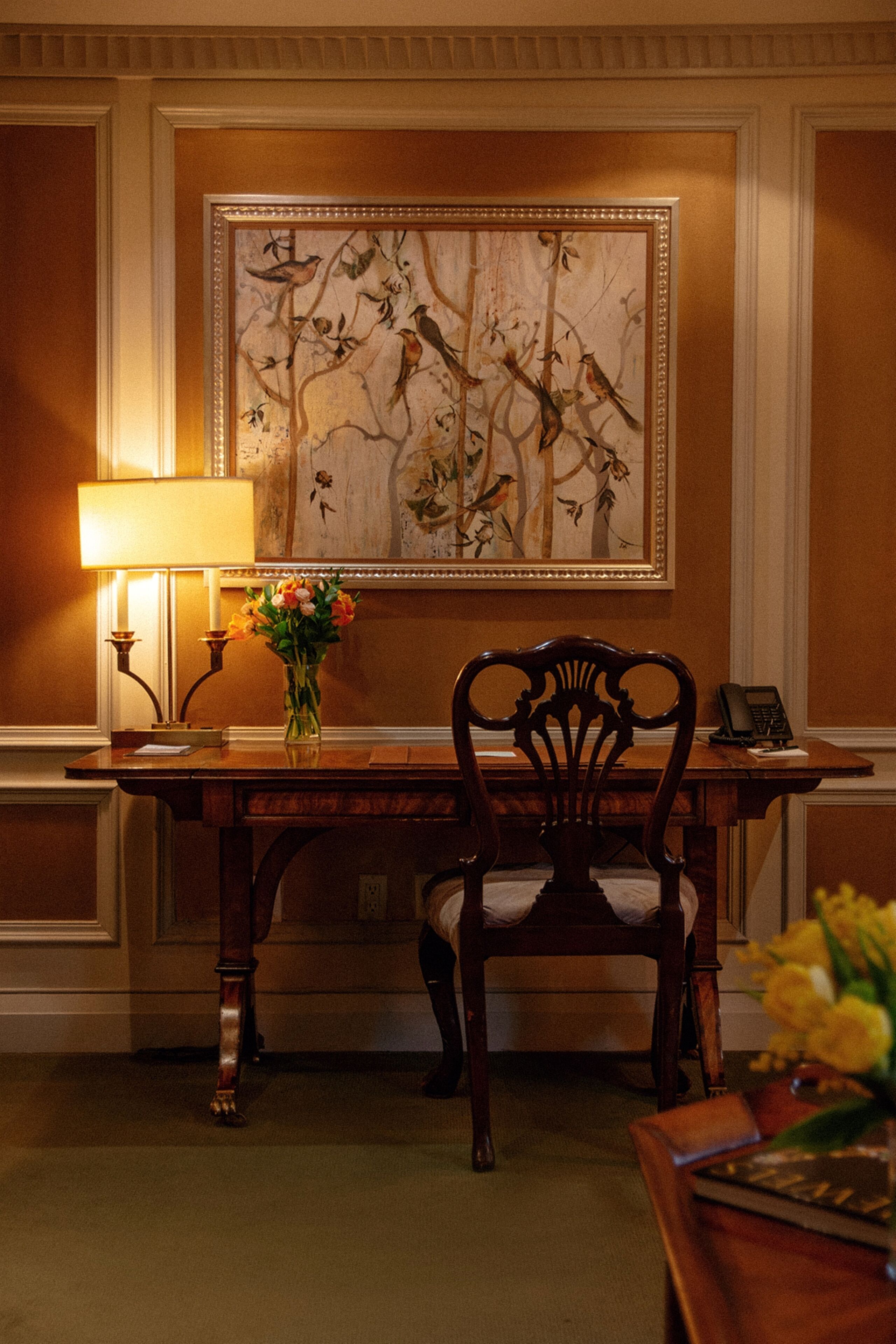 Une pièce comprenant un bureau en bois, une chaise, une lampe, un vase avec des fleurs et une œuvre d’art encadrée accrochée au mur.