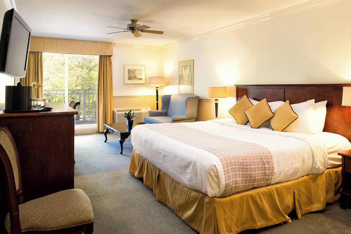 Ein Hotelzimmer mit einem großen Bett, einem Schreibtisch mit Fernseher, einem Sessel, einer Schreibtischlampe und einem Deckenventilator.