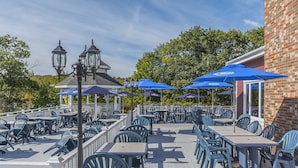 An outdoor patio with tables and chairs, blue umbrellas, and a brick building in the background. - Inn on the Lake (Fall River)