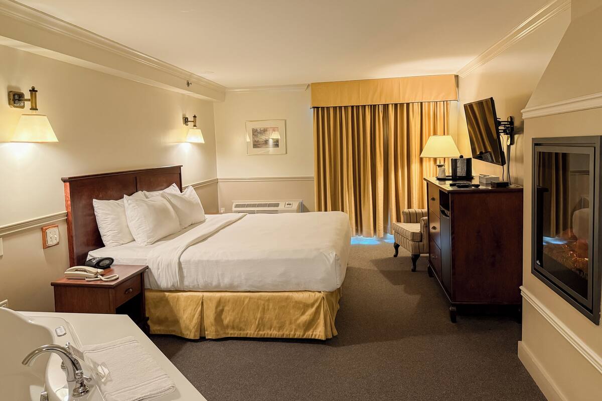 Ein Hotelzimmer mit Bett, Nachttisch mit Telefon, Schreibtisch mit Lampe, Fernseher und einem Fenster mit Vorhängen.