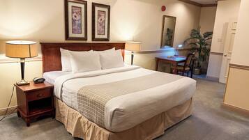 Habitación de hotel con una cama grande, dos mesitas de noche con lámparas, un cabecero de madera, un escritorio con una silla, un espejo y una planta en maceta.