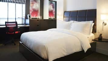 Quarto de hotel com uma cama grande, uma escrivaninha com cadeira, telefone e janela com persianas.