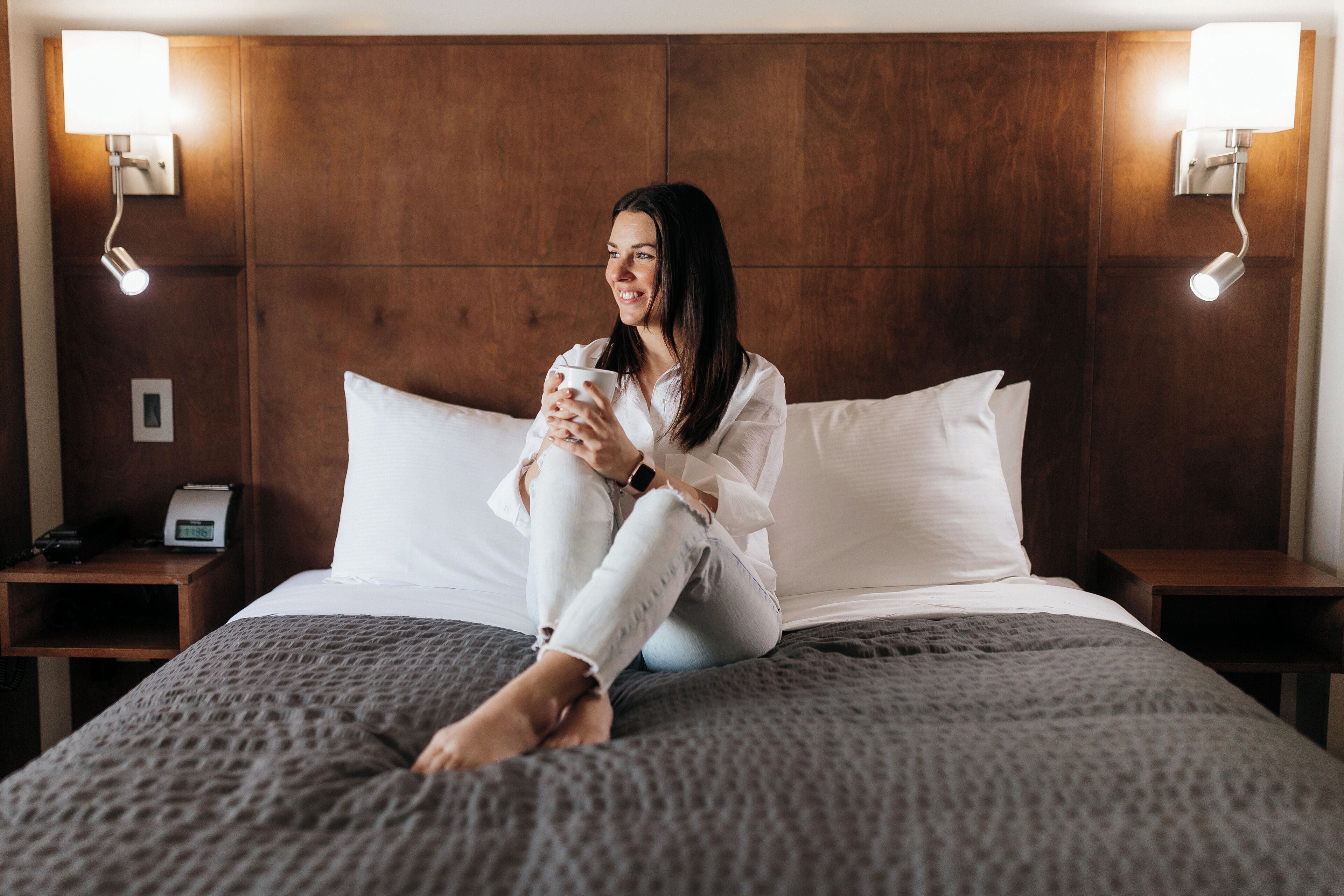 A woman sitting on a bed in a hotel room with a wooden headboard, holding a cup. There are bedside tables with lamps and a clock on one of them.