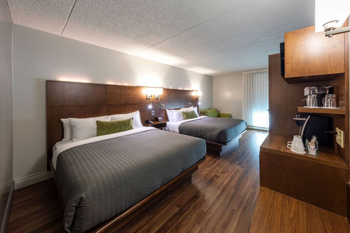 A hotel room with two beds, wooden headboards, bedside tables, a coffee maker, and a shelf with glasses.