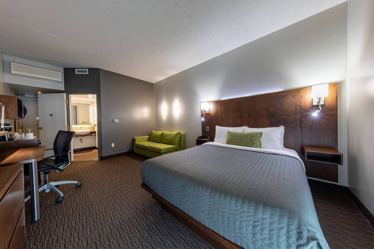 A hotel room with a large bed, a desk with a chair, a green armchair, and a bathroom visible through an open door.