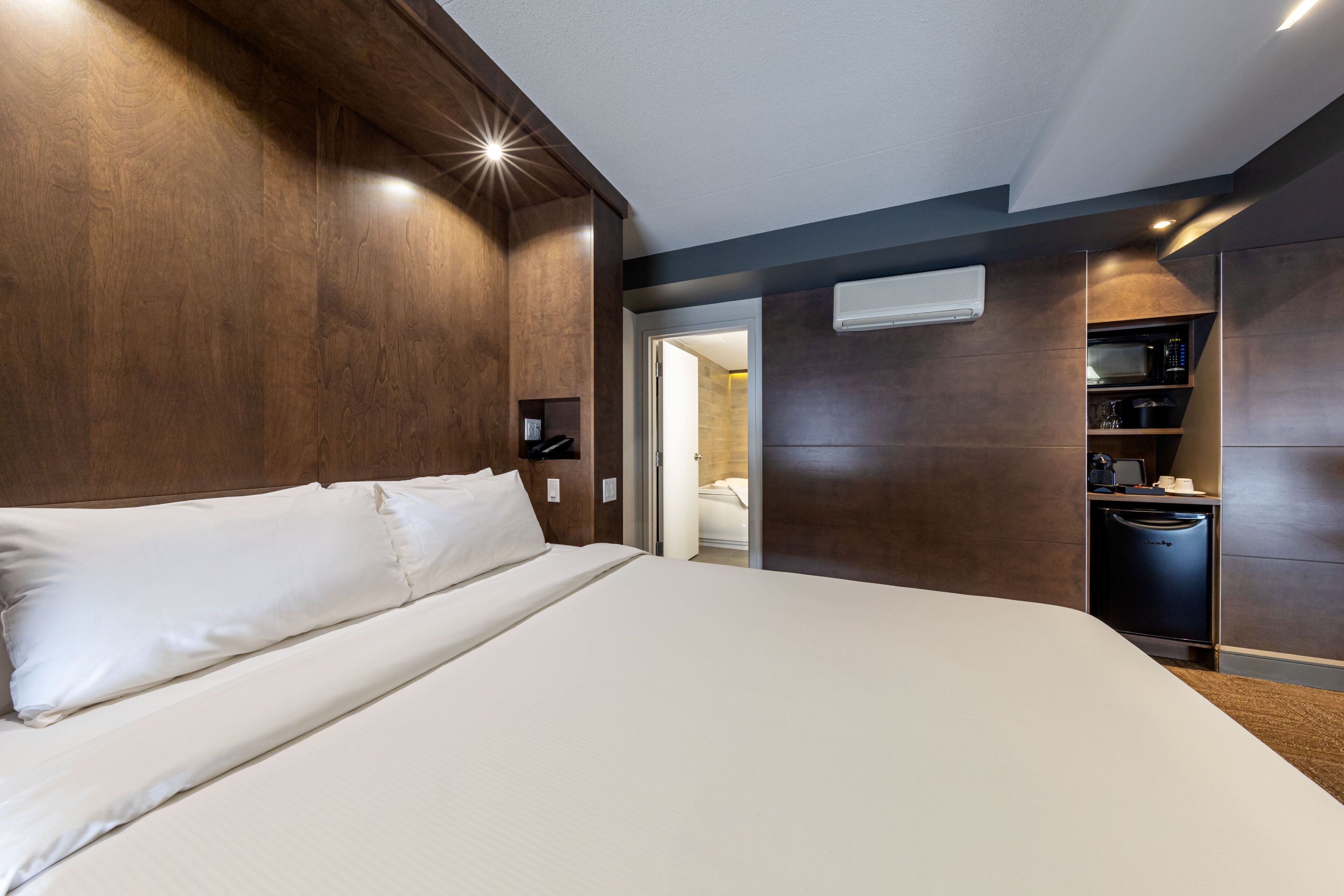 Una habitación de hotel moderna con una cama grande, cabecera de madera integrada y una cocina con microondas y cafetera.