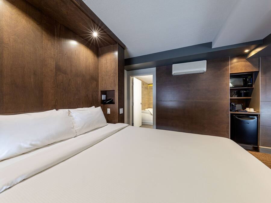 A modern hotel room with a large bed, built-in wooden headboard, and a built-in kitchen area with a microwave and coffee maker.