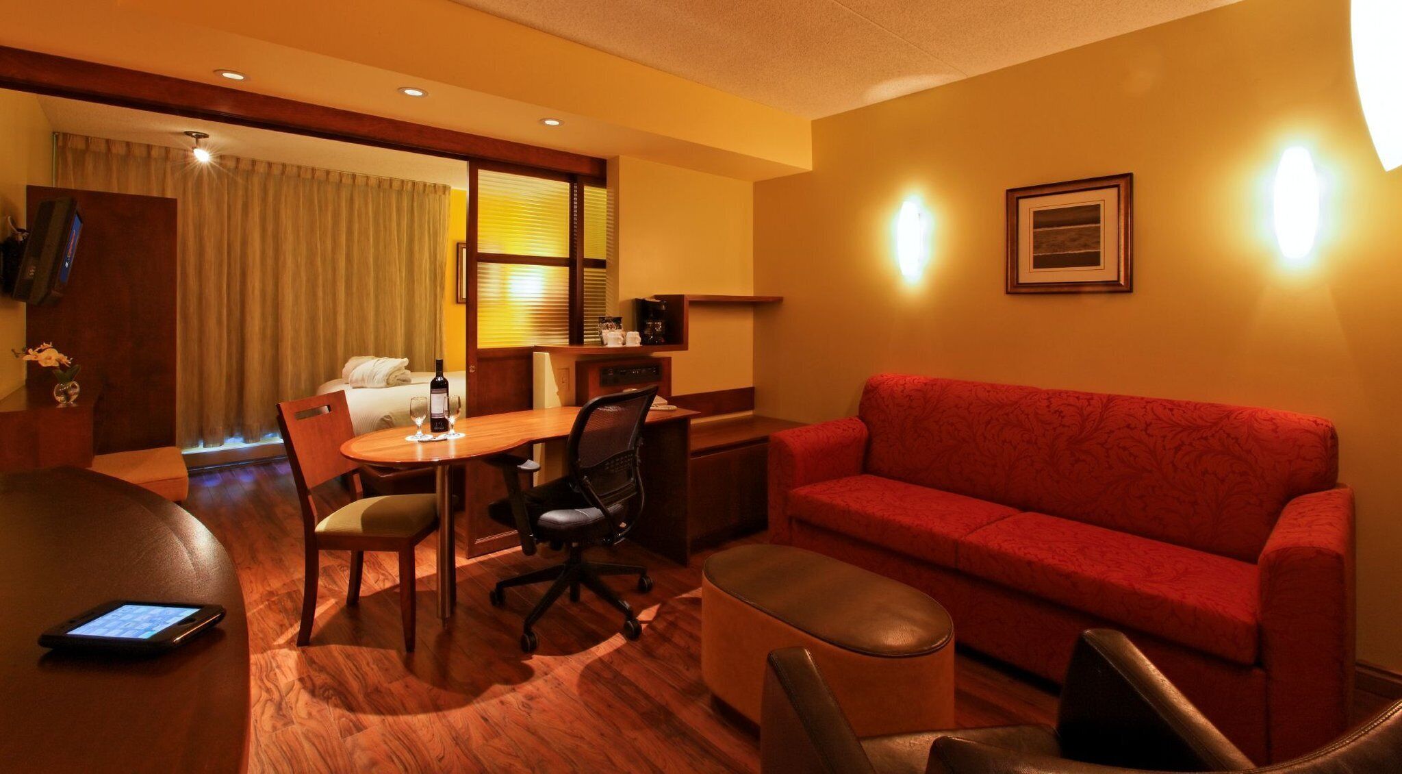 Habitación de hotel con un sofá rojo, una mesa de comedor de madera con sillas, un escritorio pequeño con una computadora, una cama con cortinas y un televisor montado en la pared.