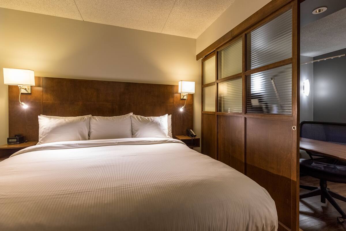 A hotel room with a large bed, two bedside lamps, a wooden headboard, a desk with a chair, and a window with blinds.