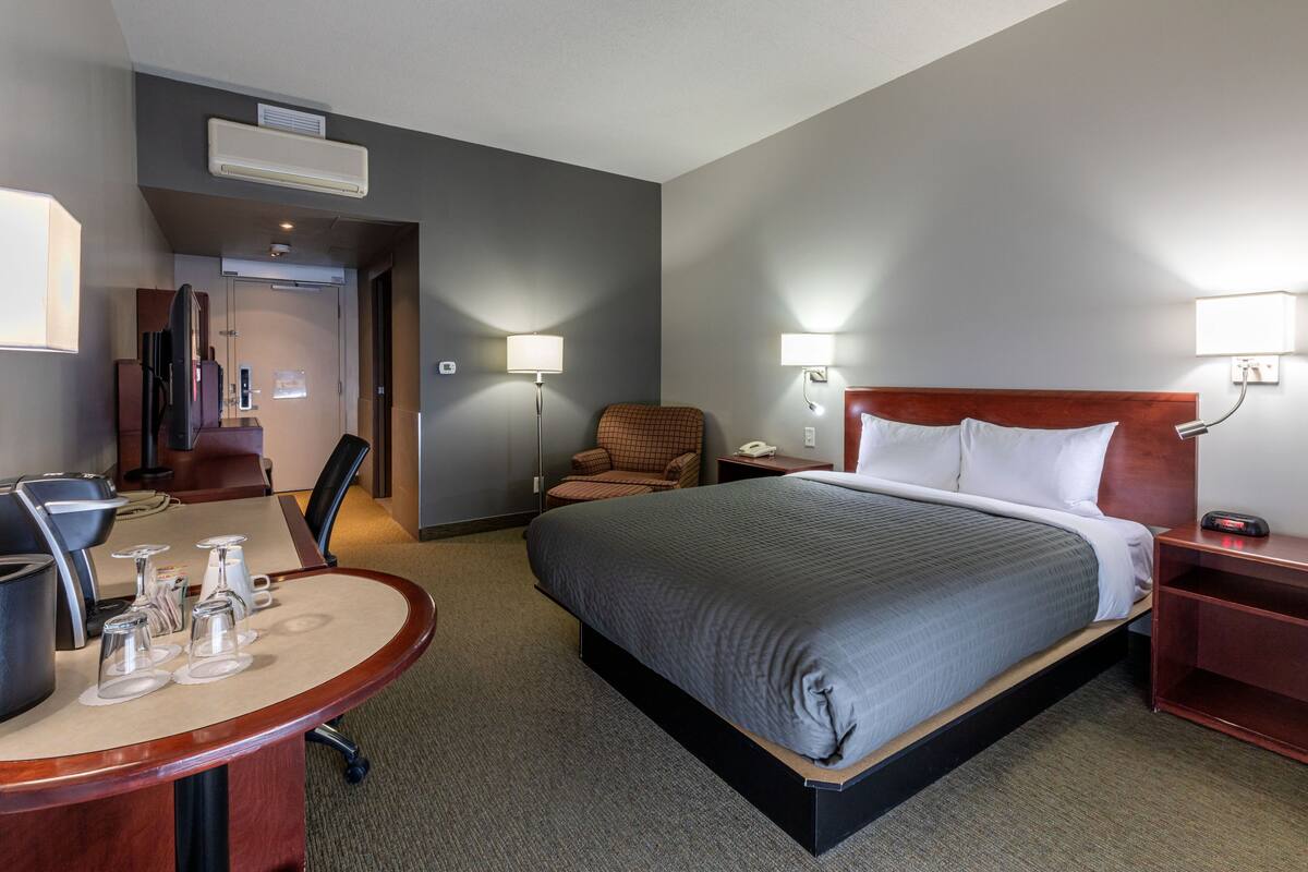 A hotel room with a bed, a desk with a chair, a chair, a nightstand, and a lamp.