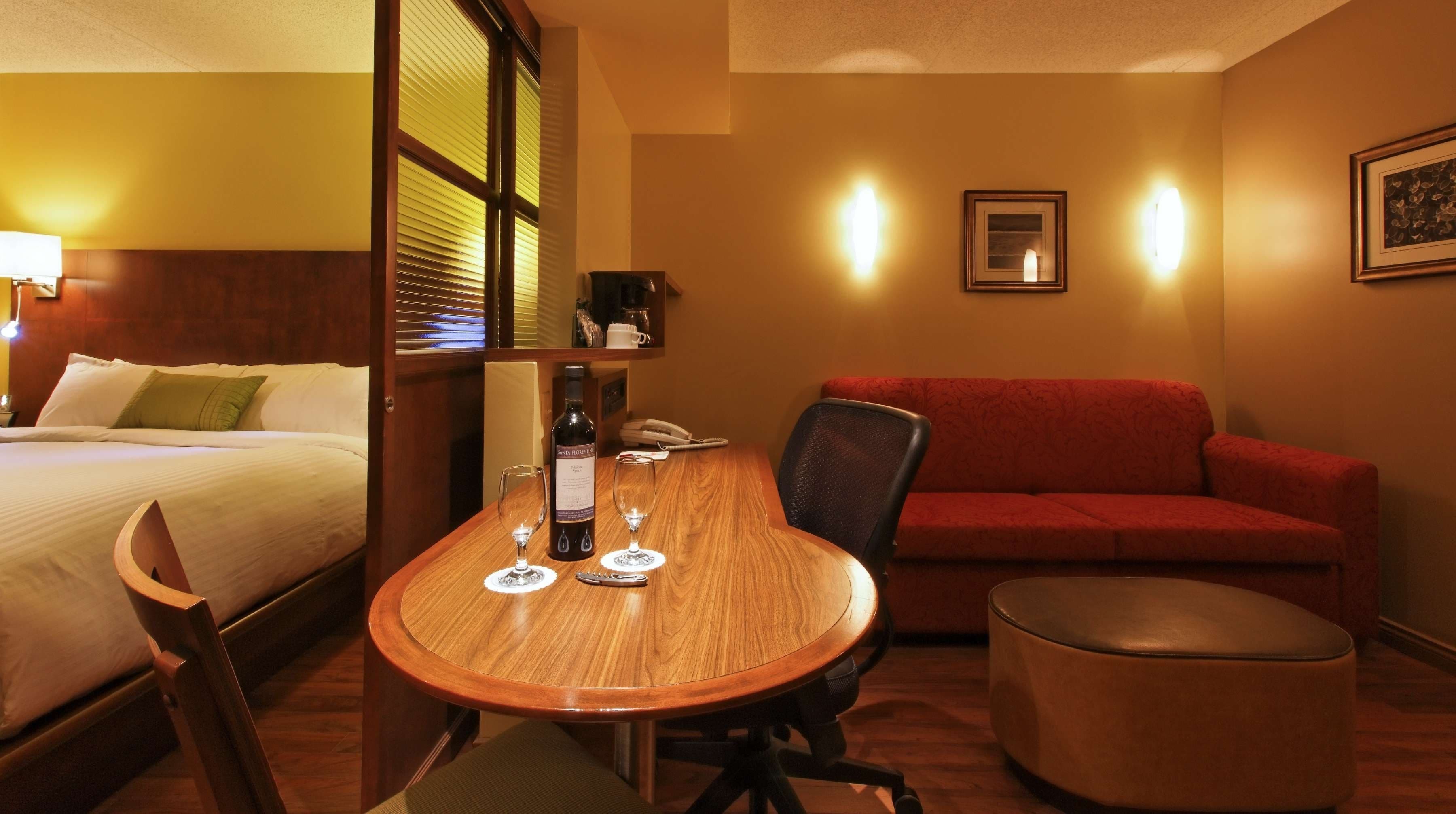 A hotel room with a bed, a wooden dining table with two wine glasses and a bottle, a red sofa, and a small ottoman.