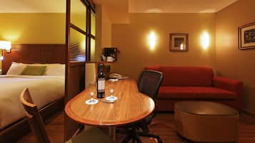 A hotel room with a bed, a wooden dining table with two wine glasses and a bottle, a red sofa, and a small ottoman.