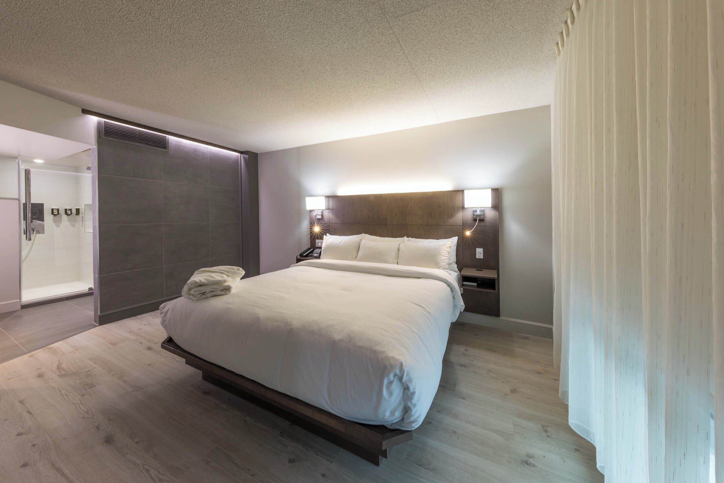 Una habitación de hotel moderna con una cama grande, mesitas de noche, ducha integrada y baño tipo suite.