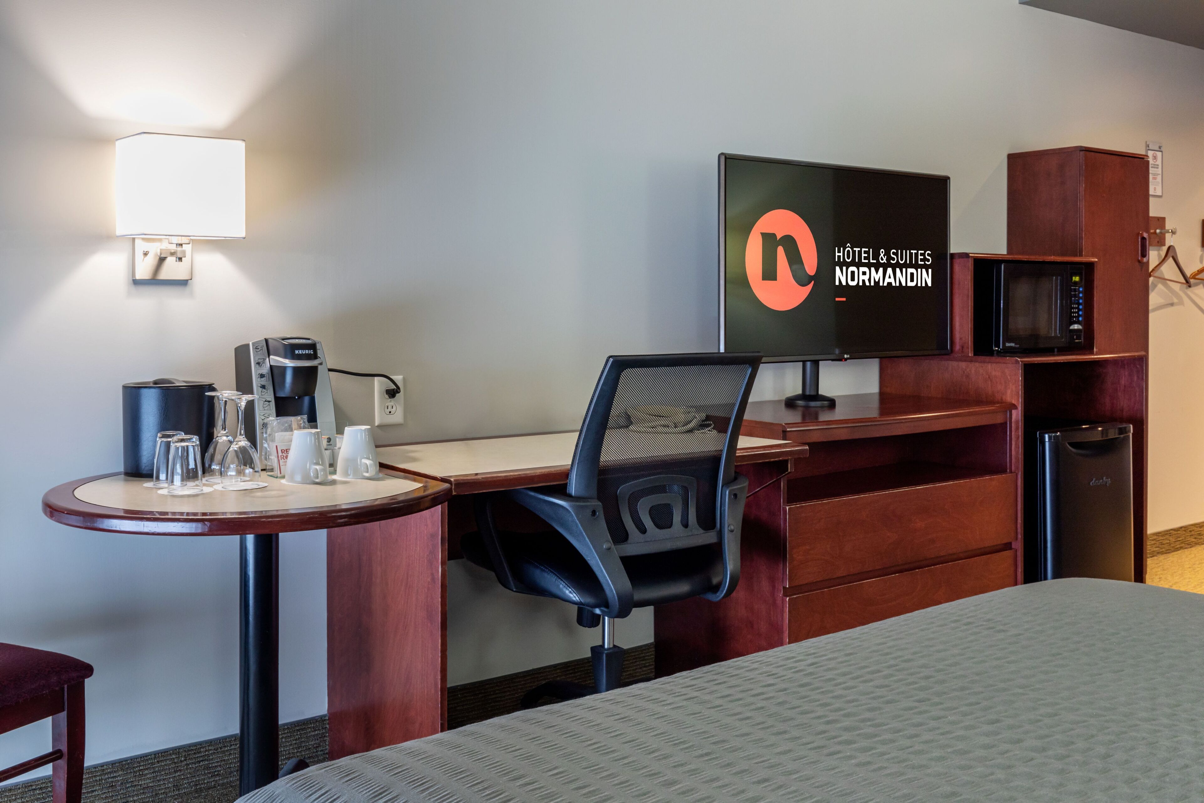 Una habitación de hotel con una cama, un escritorio con un ordenador, una silla, máquina de café, microondas y un televisor mostrando el nombre del hotel.