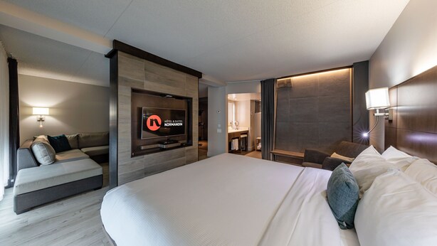 A modern hotel room with a large bed, a sofa, a TV, and a bathroom visible through an open door. - Hôtel & Suites Normandin Quebec (Québec City)