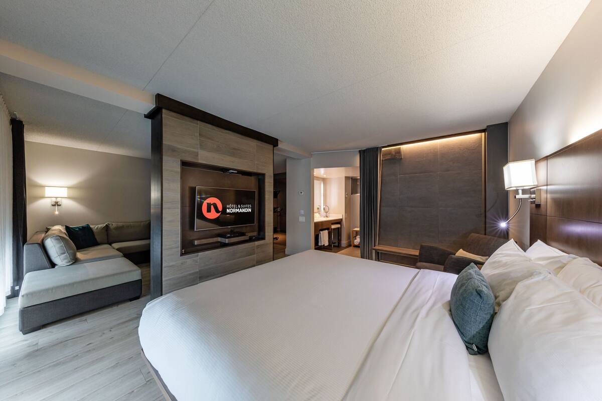 A modern hotel room with a large bed, a sofa, a TV, and a bathroom visible through an open door.