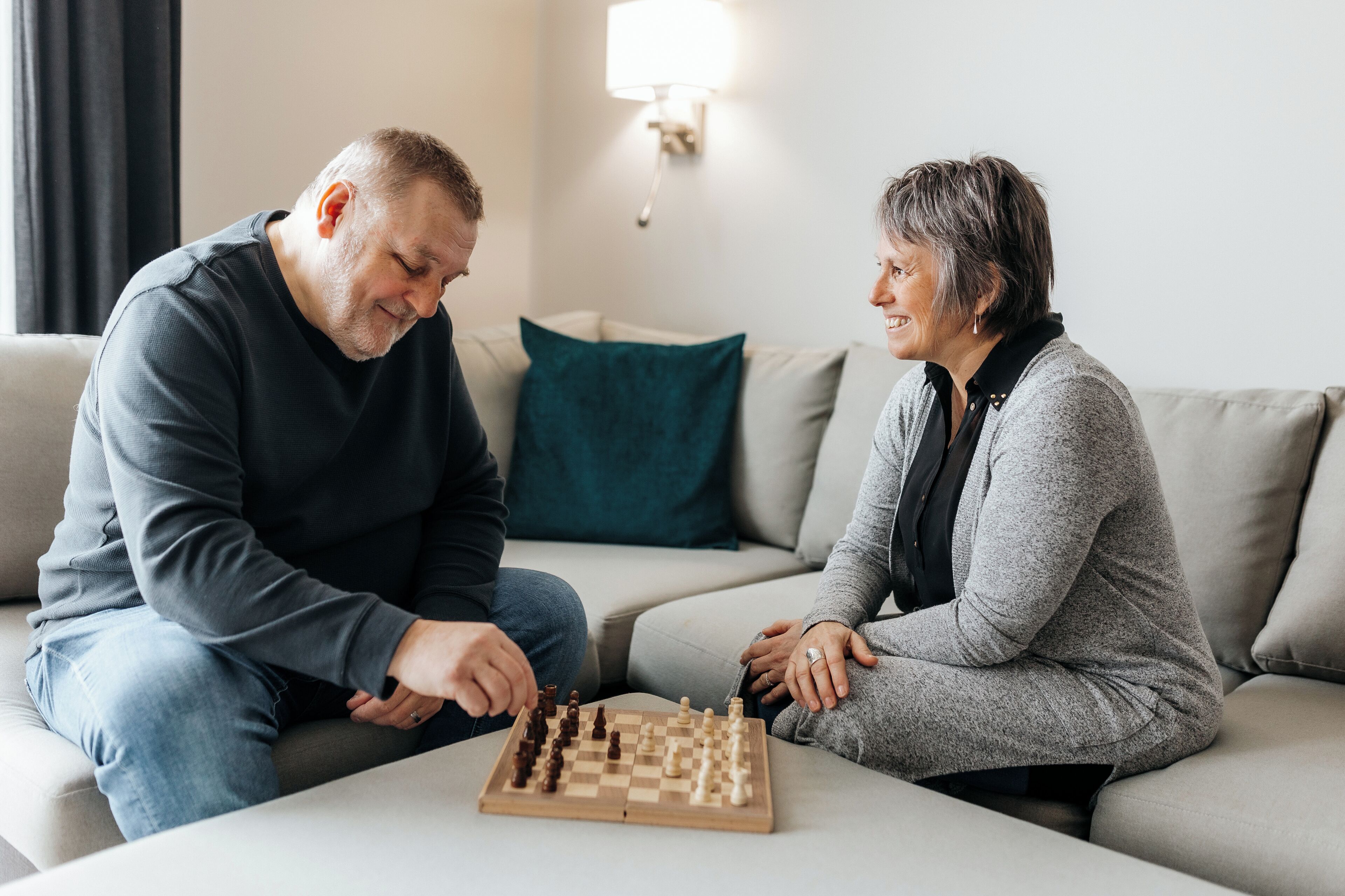 Two people playing chess on a sofa in a living room with a lamp in the background.