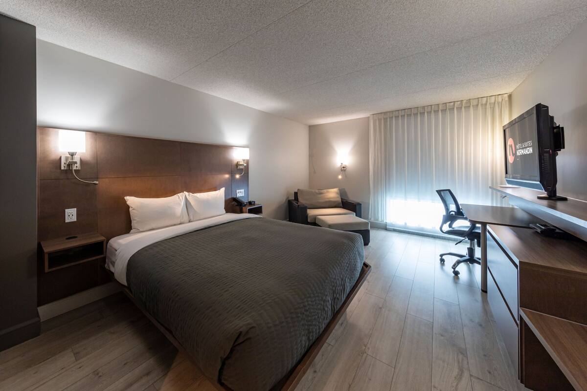 A modern hotel room with a large bed, a desk, a chair, a television, and a window with curtains.